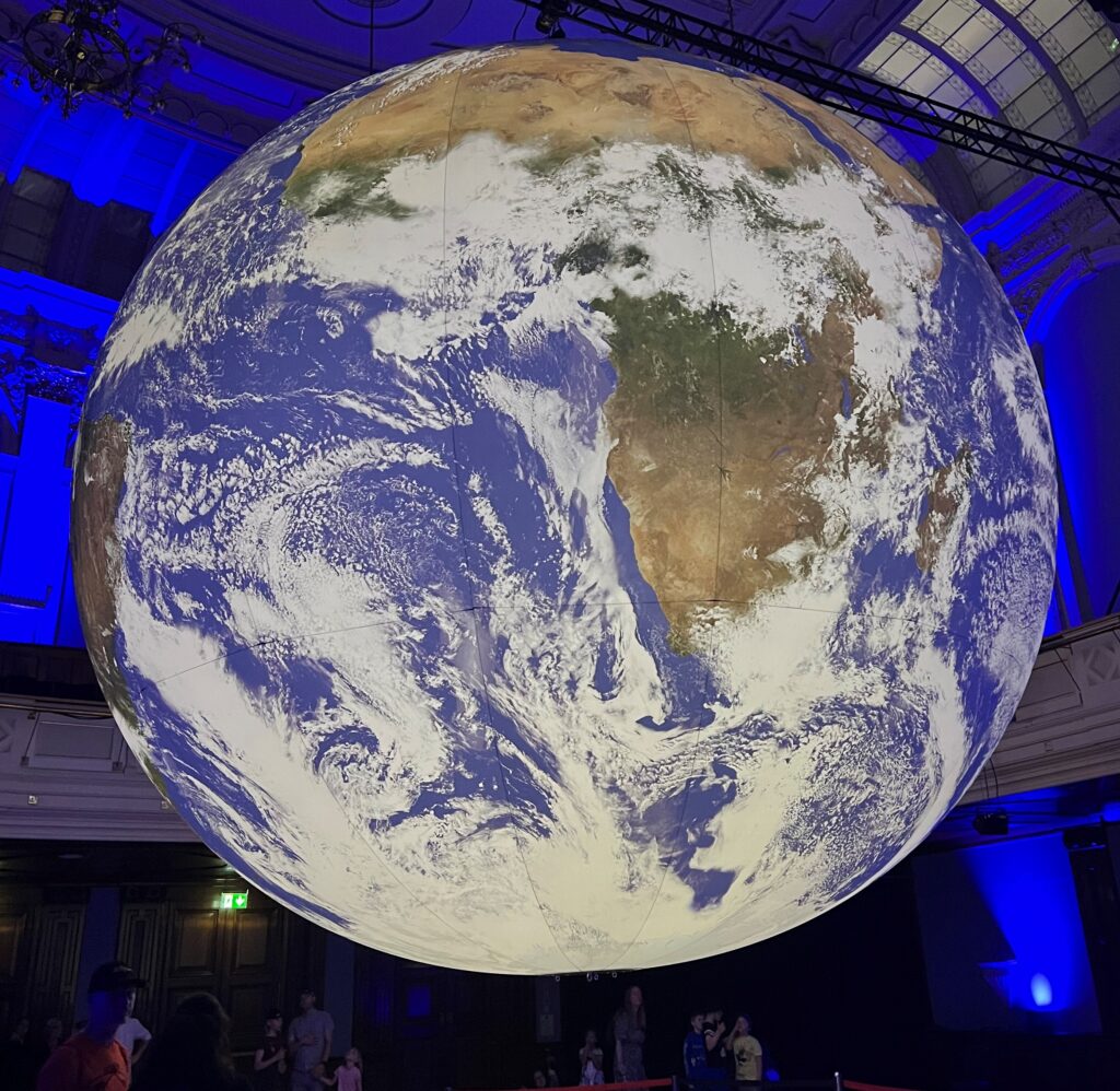 A photograph of the GAIA exhibit a 7m diameter copy of the Earth as seen from space, suspected in a concert hall.  The Earth is covered in clouds but Africa can be seen clearly sitting far higher on the globe than flat maps make us believe it is.